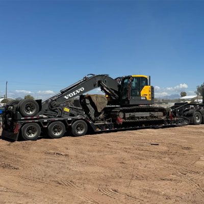 A Volvo excavator is loaded on a lowboy trailer parked on a dirt lot under a clear blue sky.
