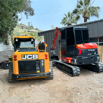 A JCB skid steer loader and a Kubota compact excavator are parked on a dirt surface near a building and palm trees.