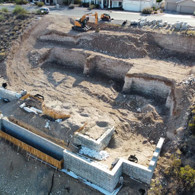 A construction site on a terraced hillside with retaining walls and excavation equipment visible, preparing land for development near residential homes.