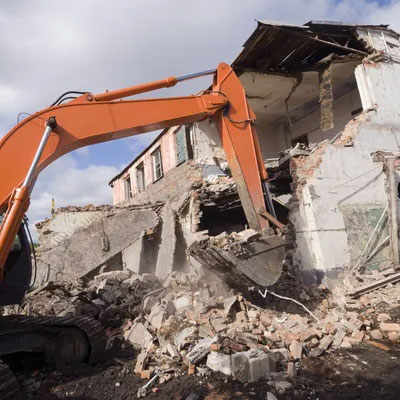 An orange excavator demolishes a partially collapsed building, surrounded by rubble and debris under a cloudy sky.