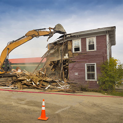 An excavator demolishes a two-story wooden house, with debris scattered on the ground and an orange traffic cone in the foreground.