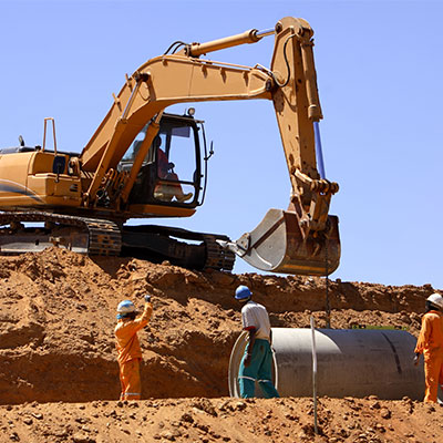 Excavator lifting a large concrete pipe while three construction workers in safety gear guide the installation at a worksite.