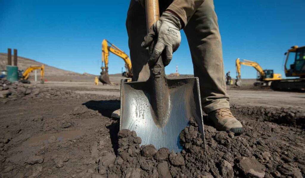 Close-up of a person digging into dirt with a shovel at a construction site, with excavators and clear blue sky in the background.