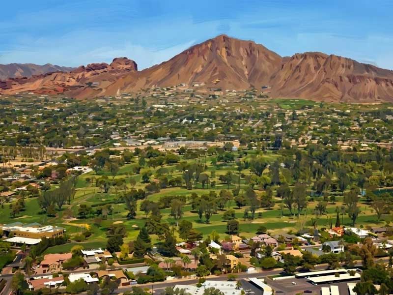 The Arcadia Phoenix AZ cityscape with scattered buildings and greenery in the foreground, with a large rocky mountain in the background under a clear blue sky.
