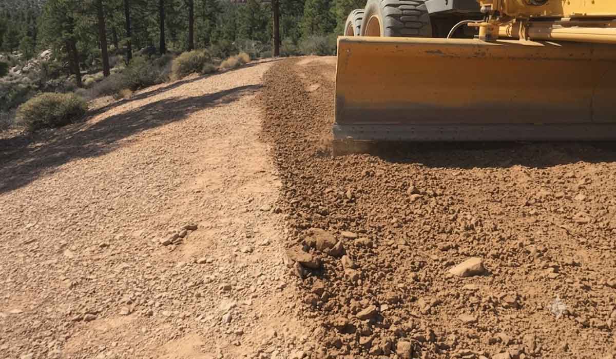 A road grader smooths and levels dirt and gravel on a rural road surrounded by trees and shrubs.